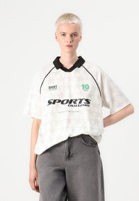 Balta marškinėliai su žvaigždžių raštu, juodas apykaklė ir žalia tekstas. Trumpomis rankovėmis, laisvas kirpimas, su užrašu "SPORTO IŠŠŪKIS" ir logotipo detalėmis.