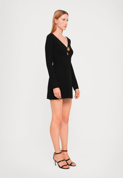 Black long-sleeve dress with a V-neck and gold floral brooch detail; fitted style and short hemline paired with black strappy high-heeled sandals.
