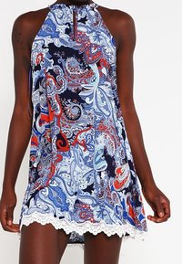 Robe sans manches avec un motif paisley bleu, rouge et blanc, décolleté halter, détail en forme de trou de serrure, et bord inférieur en dentelle blanche.