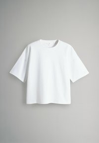 Bílý oversized tričko s krátkým rukávem a kulatým výstřihem, vystavené na jednoduchém světle šedém pozadí.