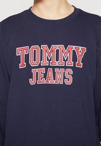 Navy blue sweatshirt with ribbed crew neck and graphic text "TOMMY JEANS" in red and white. Cotton material with a smooth texture.