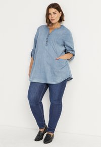 Hellblaue Denim-Tunika mit Knopfleiste und Seitentaschen. Kombiniert mit dunkelblauen, figurbetonten Jeans und schwarzen Slipschuhen.