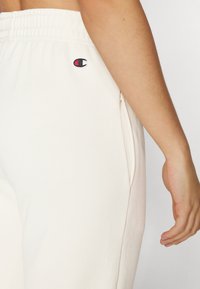 Pantalons de survêtement couleur crème avec taille élastique, poches latérales et logo brodé. Tissu doux avec une texture lisse.