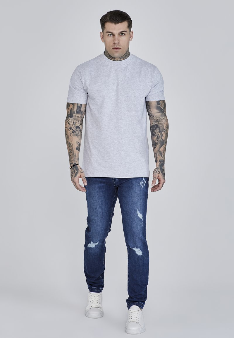 Camiseta gris de manga corta hecha de algodón, combinada con jeans azul desgastados y zapatillas blancas. El atuendo presenta tatuajes visibles en los brazos.