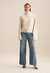 Maglione beige a collo alto con maniche lunghe abbinato a jeans a gamba larga blu chiaro e sandali neri con tacco, design minimalista e texture morbida.