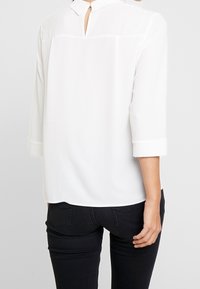Blusa bianca senza colletto con maniche a tre quarti e apertura a chiave sul retro, realizzata in tessuto morbido, abbinata a pantaloni scuri.