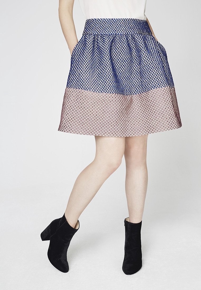 Femme portant une jupe trapèze bleu et rose à motifs avec des poches, associée à des bottines noires, debout jambes croisées.