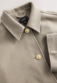 Bēšs vilnas mētelis ar struktūrētu apkakli, diviem izteiktiem zelta pogām un plāksteru kabatu. Mīksta tekstūra ar tīrām šuvēm.