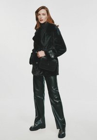 Czarny płaszcz z futra sztucznego z szerokim kołnierzem, zestawiony z błyszczącymi czarnymi skórzanymi spodniami i masywnymi czarnymi botkami. Trzyma małą czarną torbę.