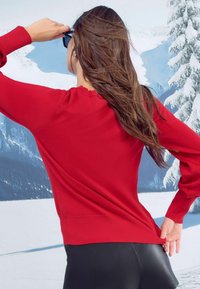 Punainen neulepusero, jossa on pitkät hihat, röyhelökaulus ja istuva muoto, yhdistettynä mustiin leggingsseihin. Taustalla on lumiset vuoret.