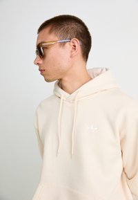 Hoodie de cor bege claro feito de tecido suave, apresentando um bolso frontal e o logótipo da adidas bordado em branco. O modelo usa óculos de sol com tonalidade castanha.