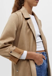 Femme portant un trench coat beige par-dessus un haut court blanc et un jean bleu, main dans la poche, sur un fond clair uni.
