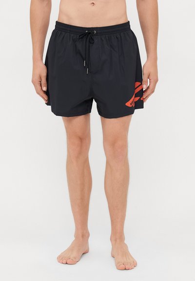 Homme portant un short de bain noir avec un graphique rouge sur le côté et un cordon de serrage, debout pieds nus sur fond blanc.