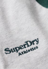 Szary materiał o miękkiej fakturze, z haftowanym tekstem w kolorze niebiesko-zielonym „SuperDry” oraz „Athletics” w celach brandingowych. Zbliżenie na wzór.