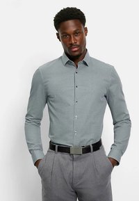 Hellblau gemustertes Hemd mit spitzem Kragen, langen Ärmeln und durchgehender Knopfleiste, kombiniert mit grauer Hose und schwarzem Gürtel.