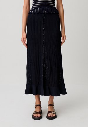 Donna che indossa una gonna midi nera in maglia con bottoni frontali e orlo arricciato, abbinata a sandali con fibbia marrone scuro.