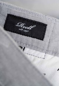 Gray corduroy fabric with a black label featuring "Reell est. 1997" in cursive. White inner lining with stitching detail visible.