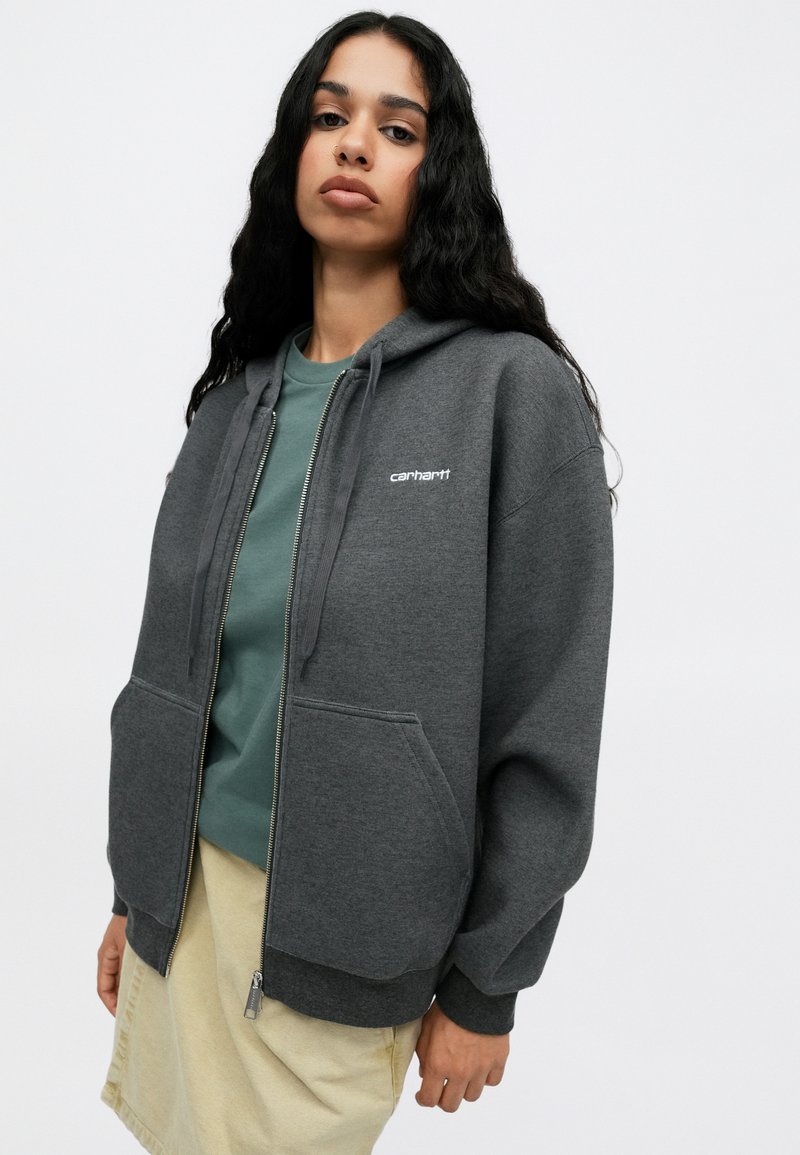 Grå zip-up hættetrøje med snøre, store frontlommer og "Carhartt" logo i hvid. Båret over en grøn skjorte og let beige nederdel.