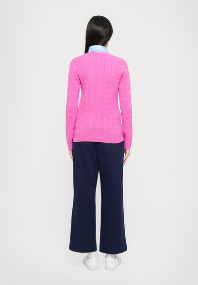 Roza pletena jopica z rebrastimi manšeti in robom, v kombinaciji z širokimi temno modrimi hlačami in belimi supergami. Zadnji pogled na obleko.