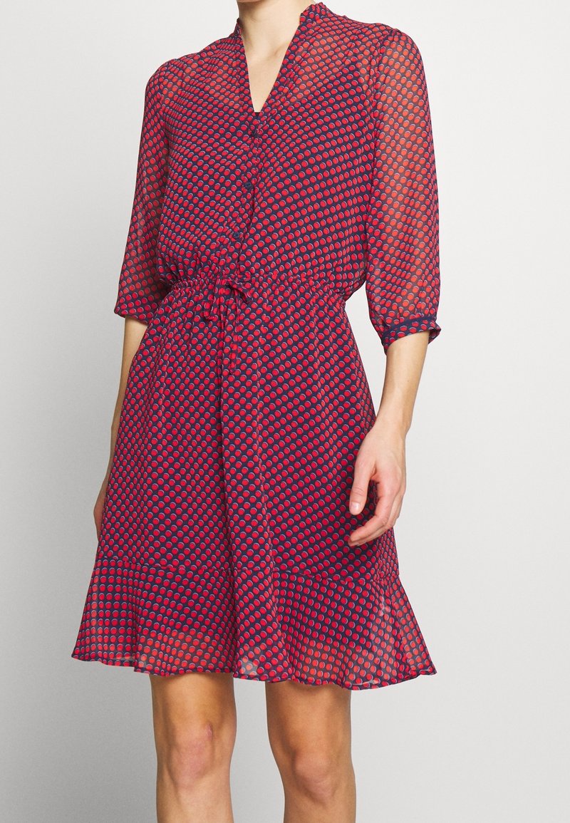 Robe rouge et bleu marine à pois, longueur genou, manches trois-quarts, boutonnée devant, ceinture à nouer et ourlet à volants.