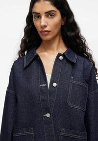 Vrouw met lang, donker golvend haar, gekleed in een donkerblauwe spijkerjack met witte knopen en beige stiksels over een lichte top.