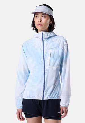 Nainen, jolla on yllään vaaleansininen ja valkoinen huppari, jossa on etuvetoketju, mustat shortsit ja vaaleanharmaa visiirillä varustettu lippalakki, katsomassa vasemmalle.