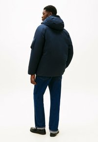 Marineblauwe pufferjas met capuchon, voorzien van een zijzak, gecombineerd met blauwe spijkerbroek en zwarte schoenen, gezien vanaf de achterkant.