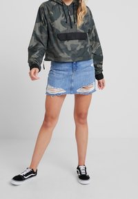Chaqueta tipo pullover de camuflaje con bolsillo frontal negro, combinada con una falda de mezclilla azul desgastada. Zapatillas negras y blancas completan el look.