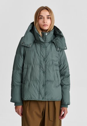 Femme portant une veste matelassée verte avec un col montant et des boutons-pression, debout devant un fond uni gris clair.