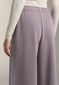Světle fialové kalhoty širokého střihu vyrobené z měkké tkaniny, s elastickým pasem a jemnými vertikálními sklady podél bočních švů.