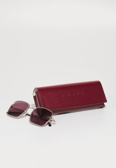 Gucci saulės akiniai su kvadratiniais, violetiniais lęšiais, juodomis rankenomis ir auksiniais akcentais, šalia bordo odinis dėklas su iškilmingu logotipu.