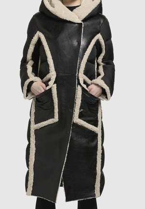 Long manteau noir avec bordure en peau de mouton beige le long des coutures, des poignets, des poches et de la capuche, porté les mains dans les poches avant.