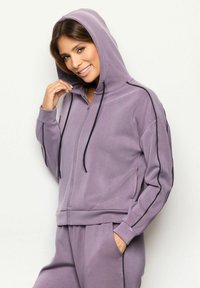 Mujer sonriendo, vestida con una sudadera con cremallera de lavanda con ribete negro y pantalones a juego, con una mano en el bolsillo, de pie contra un fondo liso.