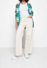 Lekkki sweter z dzianiny w kolorze teal z wielokolorowym napisem "XMAS". Biały top na ramiączkach, szerokie kremowe spodnie i białe trampki dopełniają stylizację.