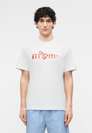 Νέος άνδρας με σγουρά μαλλιά, ντυμένος με λευκό t-shirt με κόκκινο κείμενο «msgm» και μπλε-λευκή ριγέ παντελόνι, στέκεται μπροστά σε απλό φόντο.