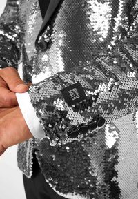Srebrna blazerka z cekinami, charakteryzująca się teksturowaną, refleksyjną powierzchnią, czarnymi guzikami oraz białym mankietem koszuli z metką marki.