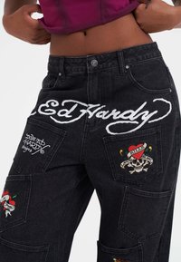 Pantalones de mezclilla negra con la marca Ed Hardy, parches bordados y varios diseños que incluyen corazones y calaveras en colores vibrantes.