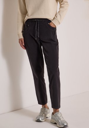 Personne portant un pantalon noir à cordon avec poches zippées, associé à des chaussures de sport argentées et un haut beige à manches longues, debout à l'intérieur.