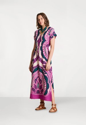 Femme debout portant une robe maxi à motifs vibrants avec des dessins roses, violets et turquoise, associée à des sandales marron et un collier bleu.