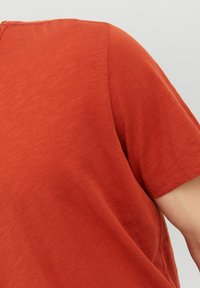 T-shirt a maniche corte in arancio bruciato, realizzato in un tessuto morbido e texturizzato con scollo rotondo e cuciture visibili sulle maniche.