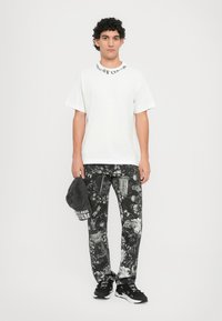 Baltas grafikos marškinėliai su juodu tekstu, juodi raštuoti kelnės su įvairiais motyvais, juoda kepurė ir juodi sportbačiai.