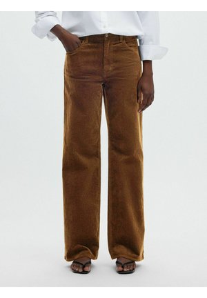 Personne portant un pantalon large en velours côtelé marron, une chemise blanche aux manches retroussées et des sandales noires ouvertes à l'avant, debout avec une main dans la poche.
