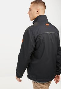 Black softshell jacket with a high collar, featuring leather accents on the shoulders and back, and a small hook detail on the yoke.