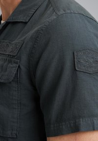 Ciemnoszara koszulka z krótkim rękawem i usztywnionym kołnierzykiem, z naszywką na lewym ramieniu i teksturowanym wykończeniem materiału.