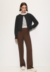 Zwarte cropped cardigan over een grijs shirt, gecombineerd met hoge bruine broek en lichte sneakers. Glad textuur overal.