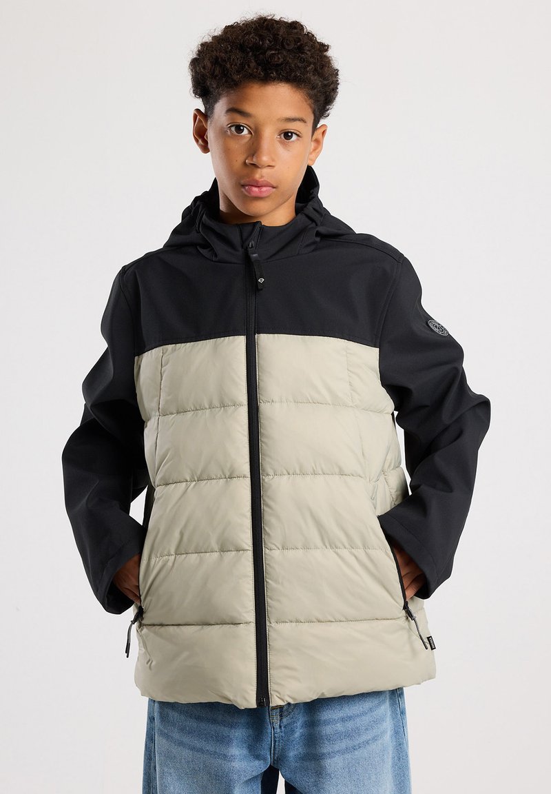 Junge Person mit lockigem Haar, die eine zweifarbige schwarze und beige Steppjacke trägt und mit den Händen in den Taschen vor einem einfarbigen hellen Hintergrund steht.