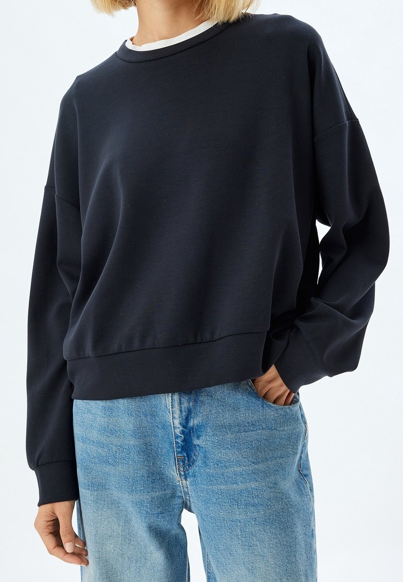 Femme portant un sweat-shirt noir à manches longues superposé sur une chemise blanche, associée à un jean bleu clair, une main dans la poche.