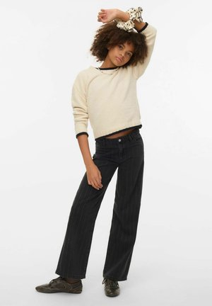 Cremefarbener, strukturierter Pullover mit schwarzem Besatz, kombiniert mit schwarzen, breit geschnittenen Jeans mit Nadelstreifen. Model trägt einen gemusterten Scrunchie am Handgelenk.