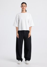 Vrouw die staat, gekleed in een losse witte korte mouw top, zwarte wijde broek en witte sneakers tegen een effen lichte achtergrond.