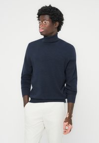 Mornarsko turtleneck pulover iz mehkega materiala, z dolgimi rokavi in ribanim robom ter ovratnikom, v kombinaciji s svetlimi hlačami, stoji pred belo ozadjem.